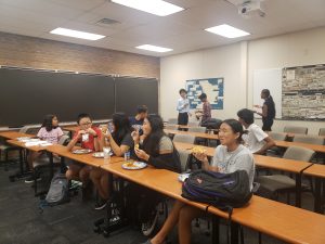 Students seated at desks in a classroom eating pizza while other people stand in the background.