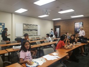 A group of students seated around a classroom eating snacks. Some students are standing and conversing. A projector and whiteboard are visible on the wall, as well as various posters.