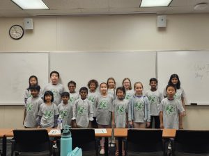 A group of children wearing matching gray shirts with a green logo, standing in front of a whiteboard in a classroom. The whiteboard has some writing on it, and a clock shows 2:42.