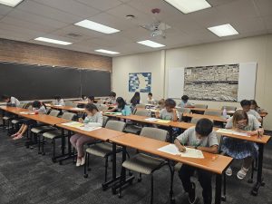 Students sitting in rows at desks in a classroom, focused on their work, with a blackboard, a bulletin board, and overhead lights.