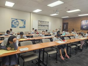 Students are seated in a classroom, working on papers at long, shared desks. The room is well-lit with ceiling lights, and educational materials are visible on the walls.