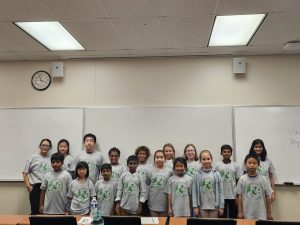 A group of children and two adults, all wearing matching t-shirts, pose for a photo in a classroom with whiteboards and a clock in the background.