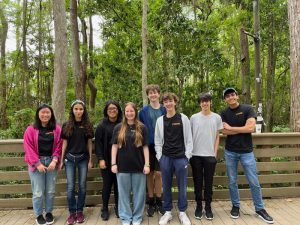 A group of nine people, standing on a wooden deck in an outdoor forested area, pose for a photo. Some are wearing black shirts with text, while others are in casual attire.