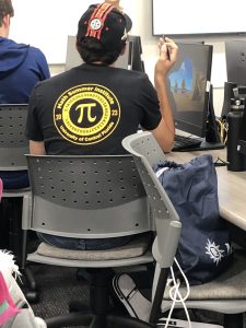 A person wearing a black hat and a black "Pi Summer Institute 2023" shirt is seated in front of a computer. They are holding a pen and a drawstring backpack is hanging on the chair.