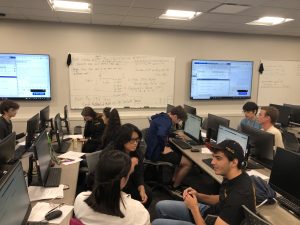 A classroom with students working on computers, whiteboards featuring handwritten notes, and large screens displaying data and graphs.