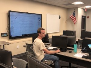 A person sits at a desk in a classroom, facing a large screen displaying a graph. The room has multiple computer stations and an American flag on the wall.