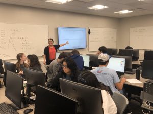 A woman points at a screen while students work at computers in a classroom. The whiteboards in the room are covered with writing and diagrams.