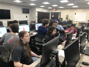 A group of students is working on computers in a classroom setting. Some students are collaborating and discussing, while others are focused on their screens. The room is equipped with multiple monitors.