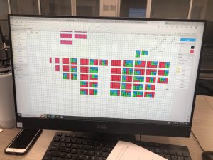 A computer monitor displays a grid-based software interface with colorful patterns. A water bottle, smartphone, paper, and pen are on the desk nearby.