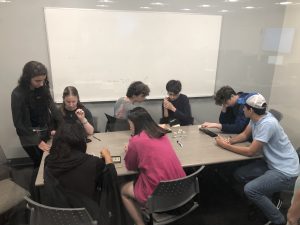 A group of eight people are seated around a table in a room with a whiteboard, engaging in a board game or activity. Two individuals are standing, observing and participating in the discussion.