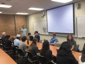 A group of people sit around a long conference table in a room with brick and white walls, a whiteboard, and a projector screen. One person stands near the screen, appearing to be speaking.