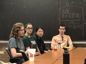 Four people sitting at a table in front of a blackboard covered with mathematical equations. They appear to be in a discussion. Various beverages are on the table.