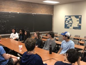 A group of students sit in a classroom with desks arranged in a U-shape, engaging in a discussion. A chalkboard with equations is visible on the left, and a bulletin board is on the right wall.