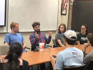 Several people are seated around a conference table in a discussion. A person with a beard in the center, wearing a plaid shirt, is gesturing with their fingers. A drink cup is on the table.