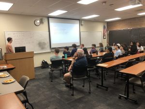 A person is presenting a slideshow to an audience in a classroom. There are notes written on whiteboards and an American flag on the wall. Several people are seated, listening attentively.