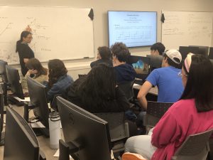 A woman stands at a whiteboard explaining a diagram to a group of students seated in a classroom. An adjacent screen displays a slide about "Converting Transition Graphs to Deterministic Finite Automata.