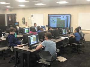 A classroom with students sitting at desks with computer monitors, engaged in activities. A large screen at the front displays a program interface. The room has rows of desks and several students present.