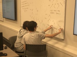 Two students collaborate at a whiteboard filled with mathematical equations and tree diagrams, pointing at various parts of the diagrams while sitting on gray chairs in a classroom.