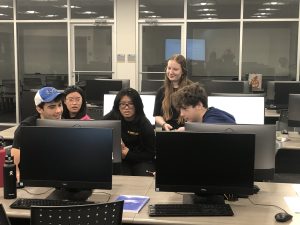 A group of five people sit and stand around computer monitors in a classroom or office setting, engaged in discussion.