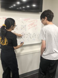 Two people are standing in front of a whiteboard covered with geometric diagrams and annotations, engaged in a discussion. One person is pointing at the board while holding a marker.