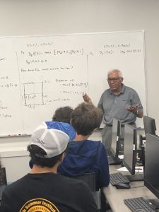 An instructor stands in front of a whiteboard filled with mathematical equations, gesturing as students seated at computers listen attentively in a classroom.