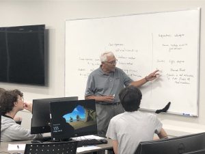 An elderly man stands in front of a whiteboard filled with mathematical notations, addressing three seated students in a classroom. Two desktop computer screens are visible in the foreground.