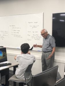 An instructor teaches a student at a whiteboard covered in mathematical equations and notes. The student sits facing the board and listening attentively.