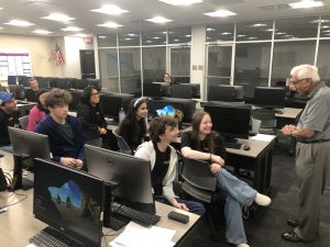 A group of people sit at computer stations in a classroom, attentively listening to an older person standing and speaking in front of them.