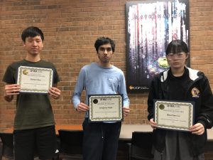 Three students standing in front of a brick wall, each holding a certificate of excellence from the WAA AMC. The certificates display their names: Daniel Zhou, Arjun Patel, and Shuchen Lu.