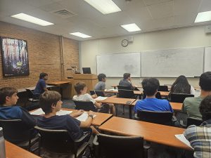 A classroom with several students seated at desks, facing a whiteboard and a lecturer at the front. The room has a brick wall, a poster, and a clock above the whiteboard.