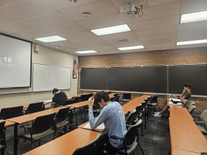 A nearly empty classroom with three students seated at desks. One student is writing, another is reading, and a third is resting their head on their hand. The room has chalkboards and an American flag.