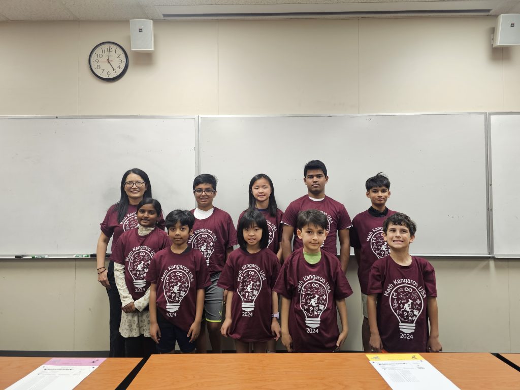 A group of children and an adult wearing matching maroon T-shirts stand in front of a whiteboard in a classroom. A clock is on the wall above them.