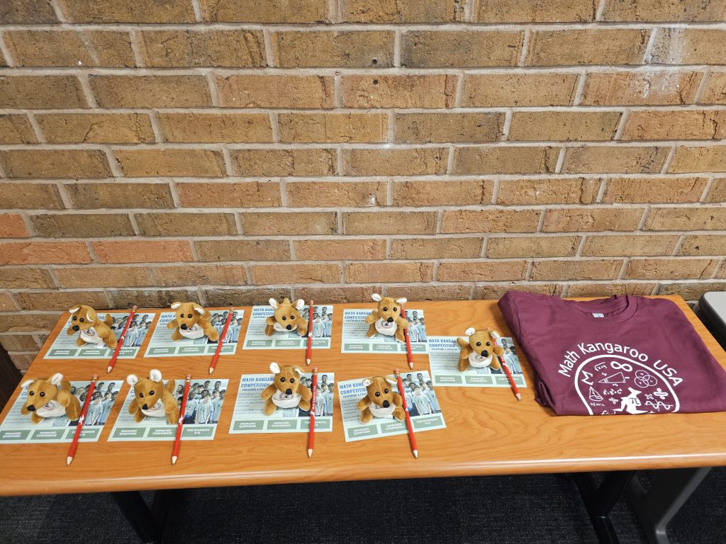 A table with informational pamphlets, pens, stuffed kangaroo toys, and a maroon "Math Kangaroo USA" t-shirt against a brick wall.