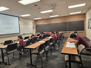 Students sit in a classroom with long tables, focusing on papers in front of them. A timer is projected on the screen at the front of the room.