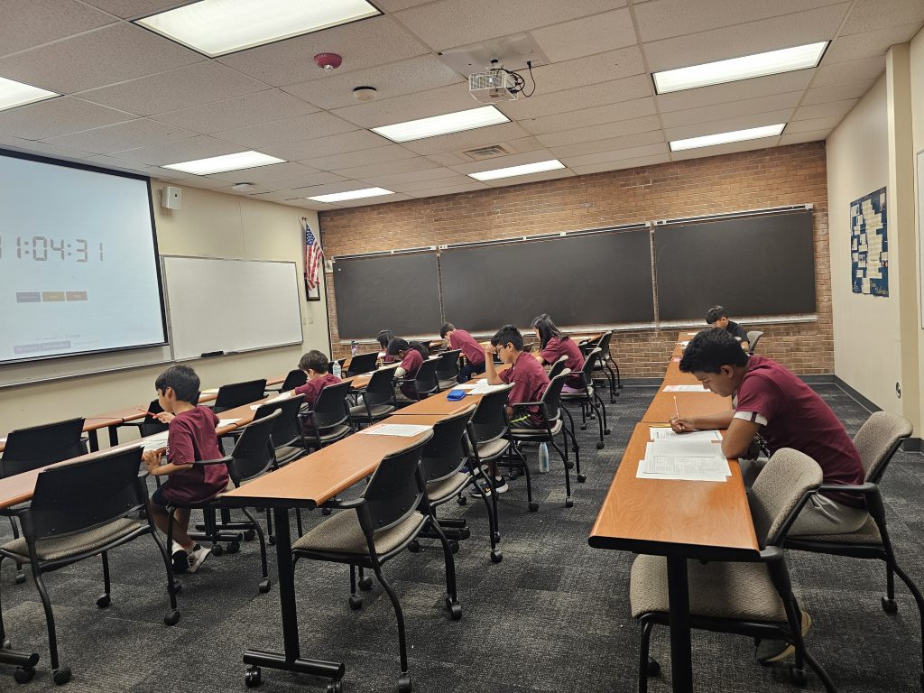 Students in matching maroon shirts are seated at desks in a classroom, each focused on a test or assignment. The room has a projector displaying a timer, a U.S. flag, and chalkboards.