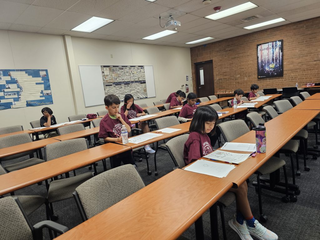 Children in a classroom sitting at desks with educational papers, engaged in writing tasks. Some desks are empty, and a large whiteboard and bulletin board are visible in the background.