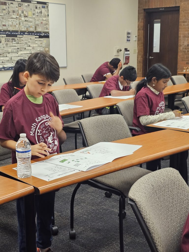 Children in a classroom participate in a math competition, each focused on their test papers. They are wearing maroon t-shirts with "Math Kangaroo" printed on them.