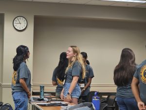 People standing and conversing in a classroom with a clock showing 11:55 AM. Some are wearing matching gray t-shirts. A table with various items is in the foreground.