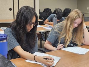 Two people sit at a table in a classroom, focusing on papers in front of them. Both are writing with pens, and there are water bottles and additional papers on the table.