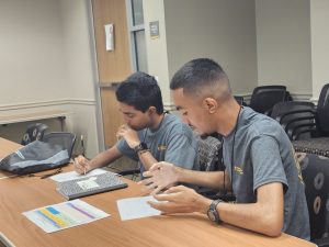 Two individuals in gray shirts are sitting at a table in a classroom, engaged in a discussion. One is taking notes while the other is gesturing with their hands. Papers and a notebook are on the table.