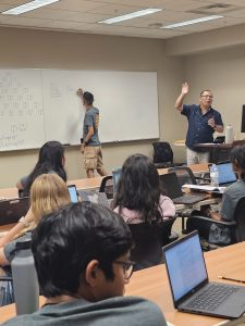 A teacher lectures to a classroom of students, one of whom is writing on a whiteboard. Other students are seated, looking at laptops and notebooks.