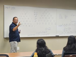 A man stands at the front of a classroom pointing to a whiteboard filled with mathematical equations while three students seated in front of him listen attentively.