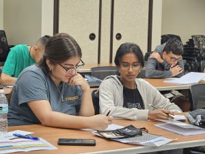 A group of students sitting at a table in a classroom, focusing on their work. Some are writing, and there are books, papers, a water bottle, and a smartphone on the table.