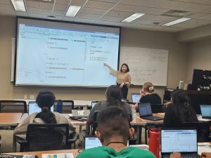 A woman stands at the front of a classroom pointing at a projected screen displaying code. Several students sit at desks with laptops, engaging with the lesson.