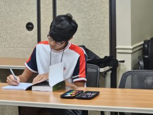 A student wearing a red and white t-shirt is writing in a notebook while looking at a textbook. Two pencil cases and a backpack are on the desk next to him in a classroom setting.