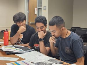 Three young men sit at a table, deeply engaged with papers and a laptop in front of them. One holds a pen to his mouth, suggesting they are working on a project or studying together.