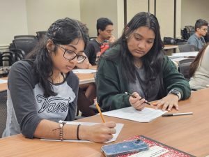 Two students in a classroom are focused on writing in their notebooks; one wears glasses and a "Vans" shirt, the other is dressed in a green jacket. Additional students are visible in the background.