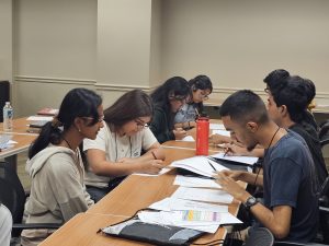 Students sitting at a table in a classroom, engaged in group study with papers and textbooks spread out. Some are writing or reading while others discuss.