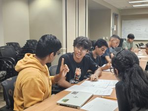 A group of students is seated at a long table, engaged in discussion and writing on papers. One student wearing a yellow hoodie is gesturing while talking to another student wearing glasses.