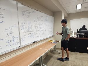 A person stands in front of a whiteboard covered in mathematical equations, holding a marker, in a classroom setting.